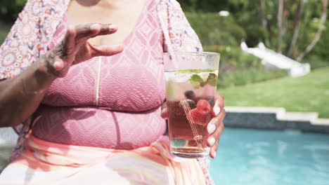 Feliz-Anciana-Afroamericana-Con-Gafas-De-Sol-Junto-A-La-Piscina-Bebiendo-Cócteles,-Cámara-Lenta