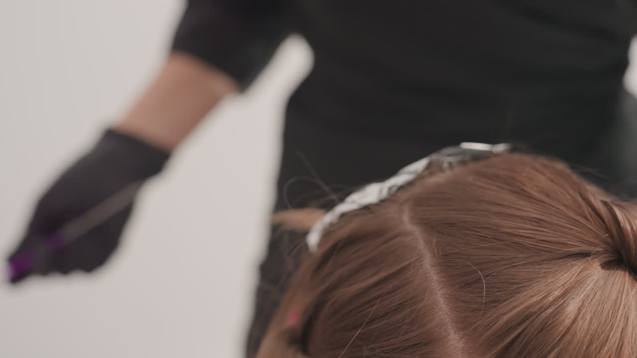 Parrucchiere che applica colpi di luce con stagnola sui capelli, mani con guanti che usano un pettine viola per dividere i capelli castani in sezioni, fogli di stagnola piegati per maggiore precisione, applicazione ravvicinata alla radice, sfondo del salone sfocato, professionale
