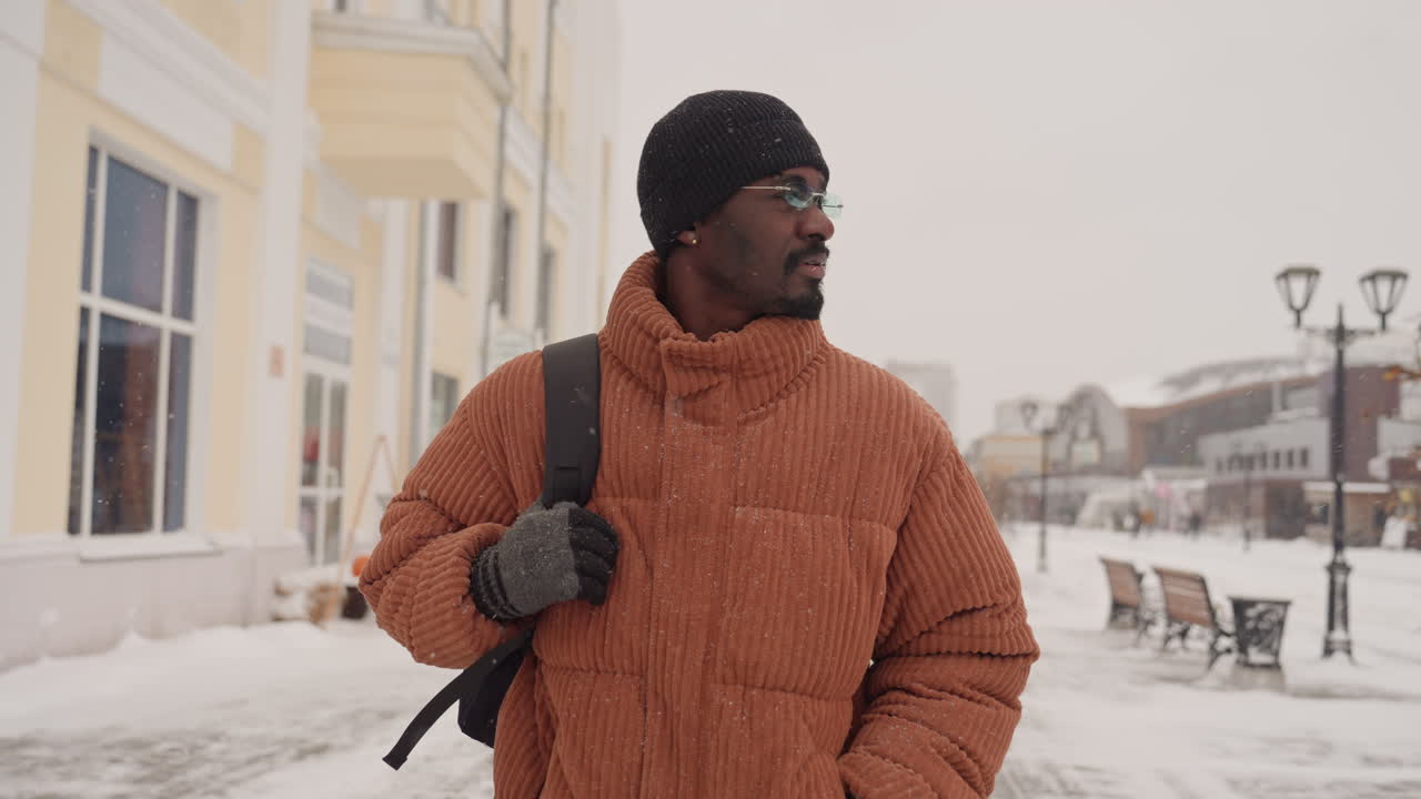 Homme noir marchant dans une rue enneigée du centre-ville, portant un épais manteau orange, un bonnet noir et des lunettes, avec un sac à dos, passant devant des vitrines et des bancs aux couleurs pastel, flocons de neige tombant, silhouettes de lampadaires.