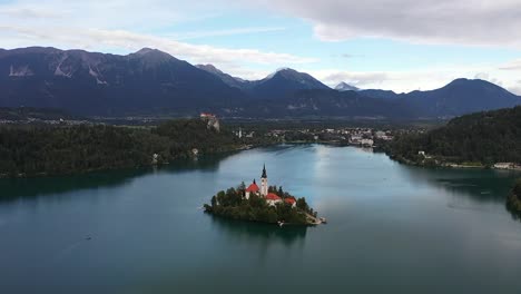 a drone slowly retreats from bled island, unveiling the fairytale church as shimmering blue waters widen and slovenia’s majestic mountains rise beyond, capturing the iconic beauty