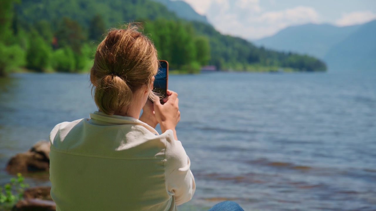 Woman sits by calm lake, phone raised to capture scenic view. She focuses intently, enjoying peaceful natural surroundings. Mountains frame water, green trees line shores. Perfect for travel, wellness