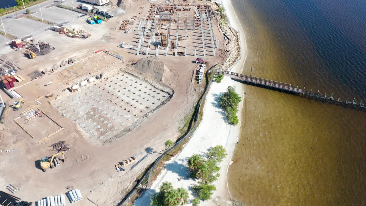 Há obras em andamento perto de uma praia de areia adjacente a um estacionamento com carros e quadras de tênis visíveis, situada ao lado de uma orla calma em um ambiente ensolarado.