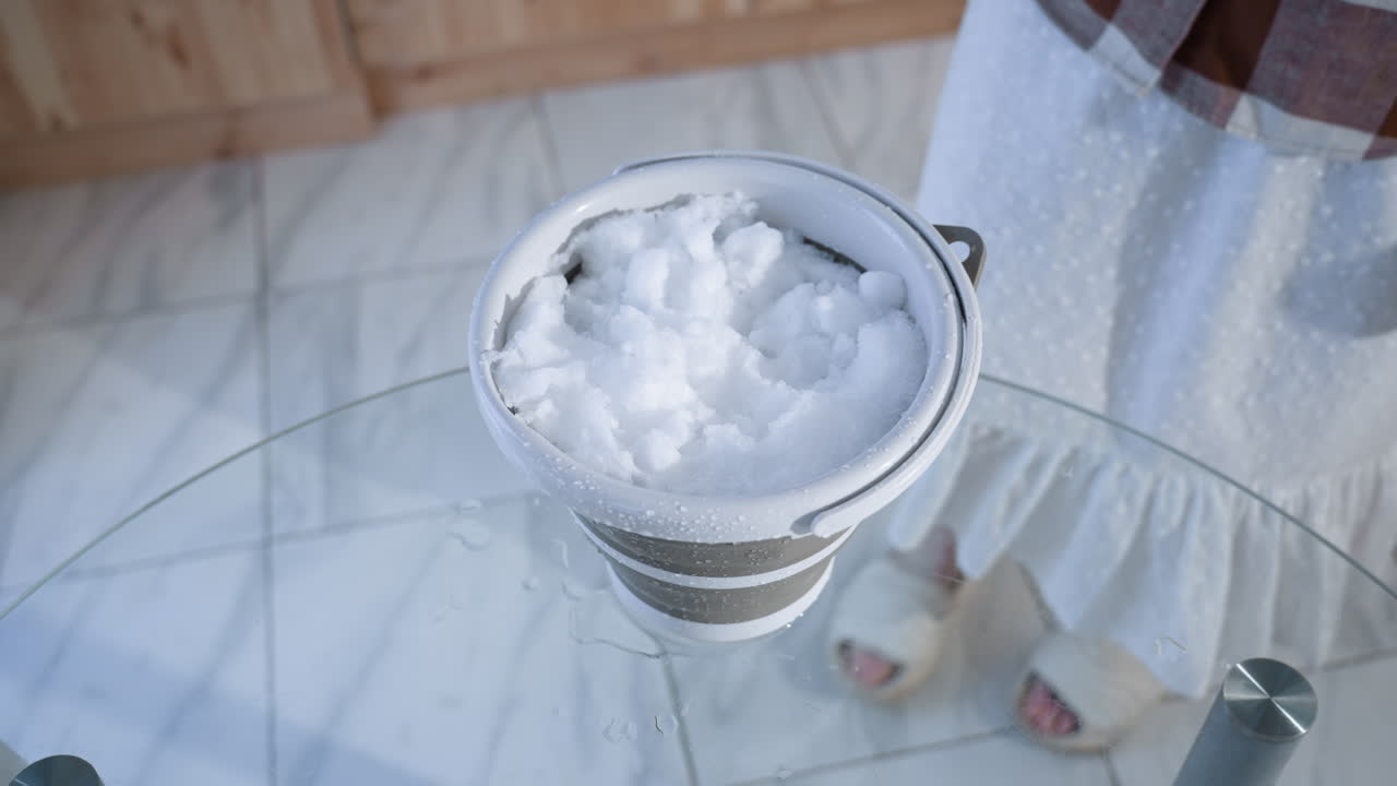 High angle view of young woman placing bucket filled with fresh snow onto transparent glass table then stepping back as water drips from melting ice, bright interior setting reflecting winter ritual