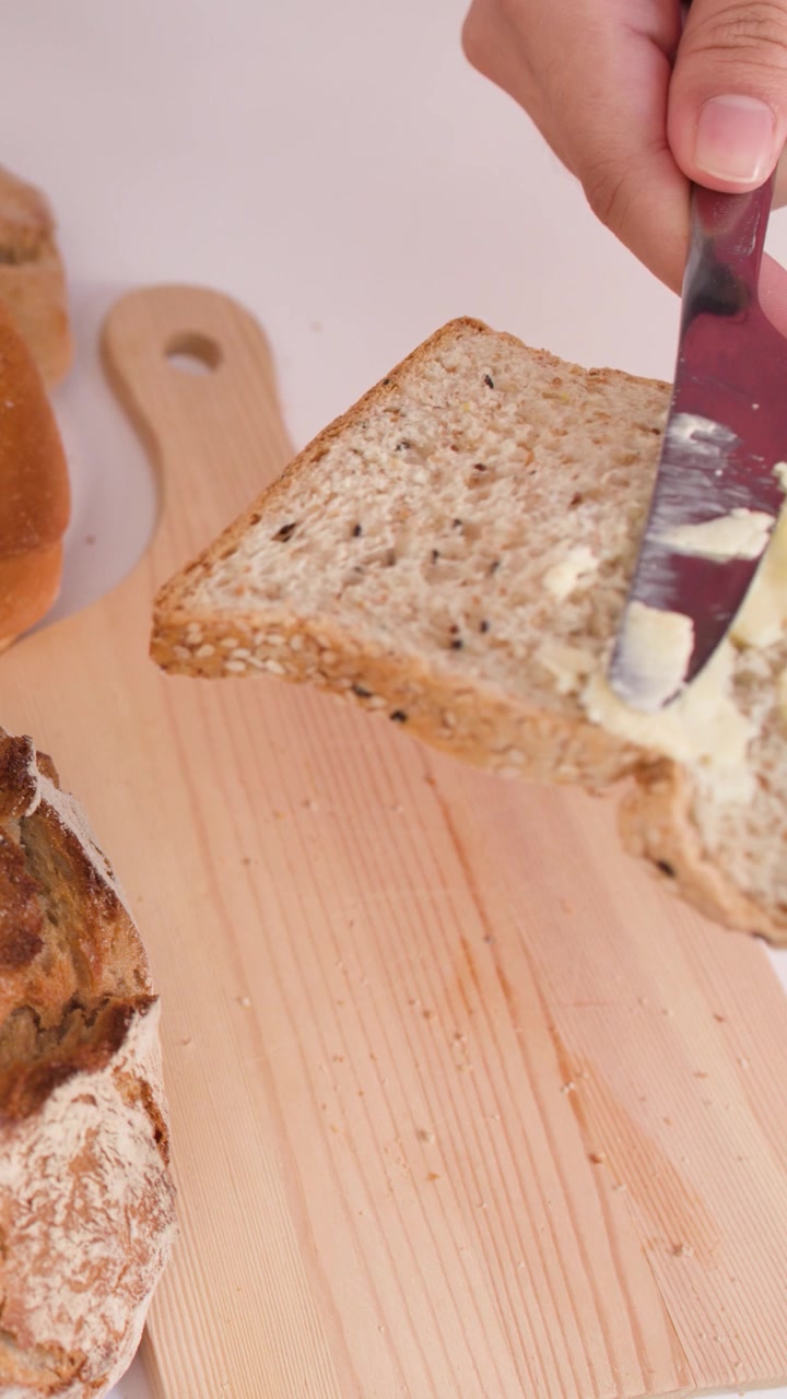Hand uses a knife to spread peanut butter on multigrain bread over a wooden board.