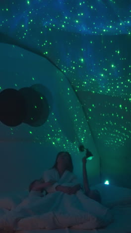 woman relaxes on comfortable bed with little kids looking at stars displayed by beamer on wall. children get ready for bed in room with subdued lighting