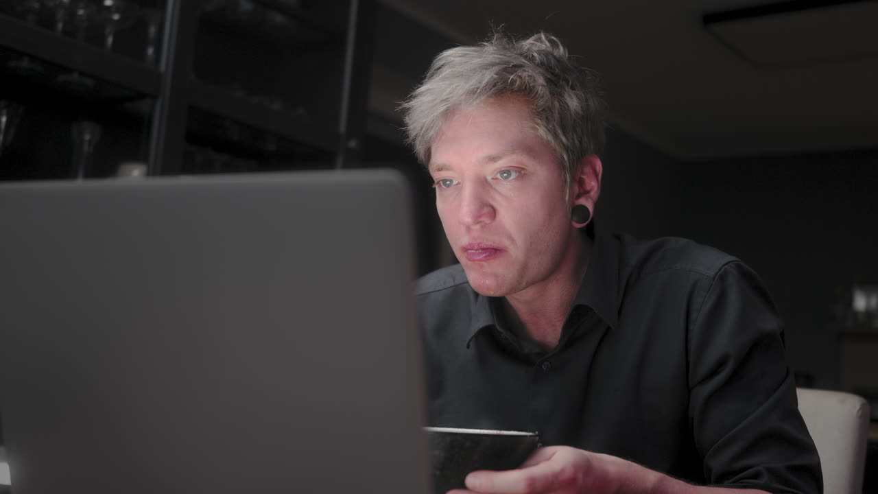Young man with silver hair and a black business shirt sitting lonely in a dark room eating asian noodle soup andwiping his mouth, while staring into a bright notebook screen