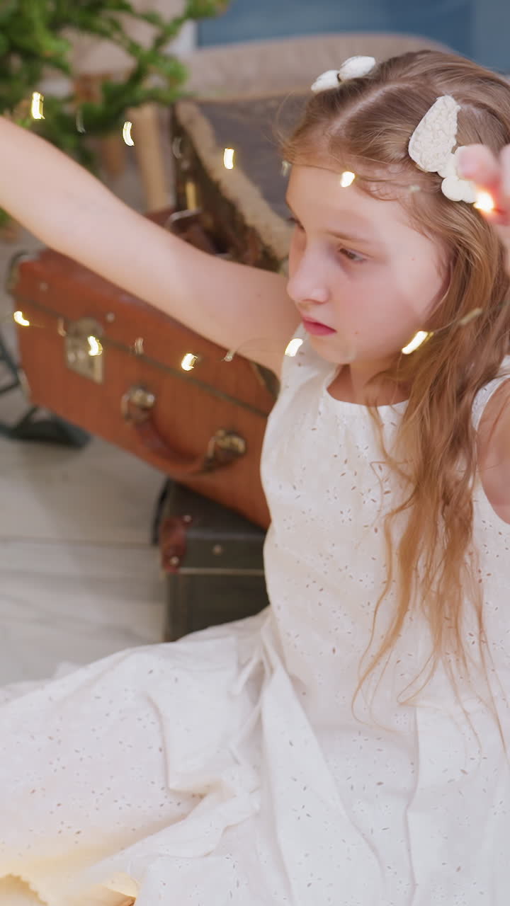 Chica blanca con luces de Navidad en interiores, sentada cerca de una maleta vintage, manipulando luces brillantes, suaves destellos dorados a la deriva, con un vestido blanco con lazo de mariposa, expresión suave y concentrada