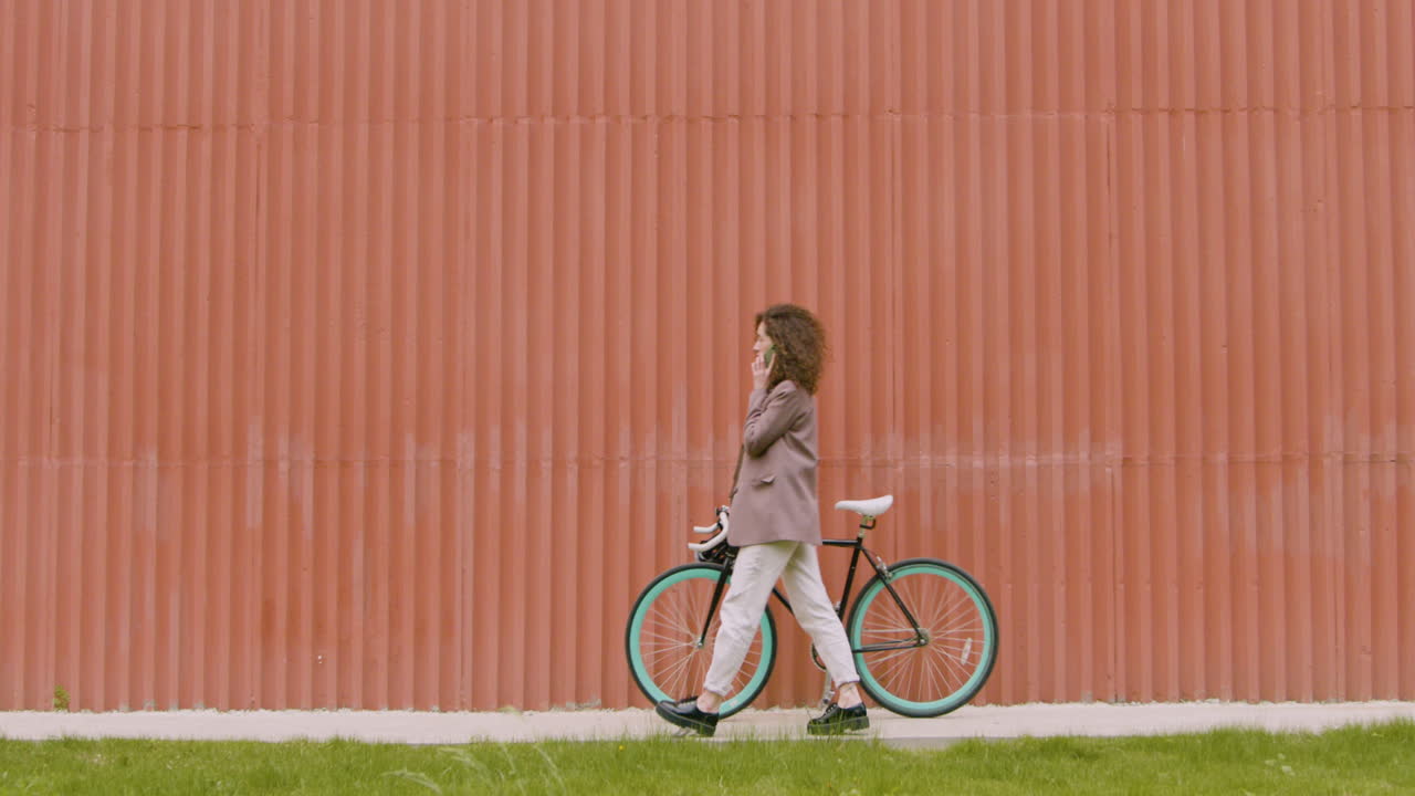 정장 차림의 젊은 여성이 자전거를 타고 걸어다니며 조립식 금속 건물 앞에서 전화를 걸고 있다