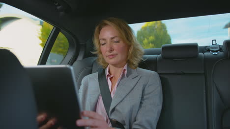 mature businesswoman sitting in back seat of taxi or car working on digital tablet