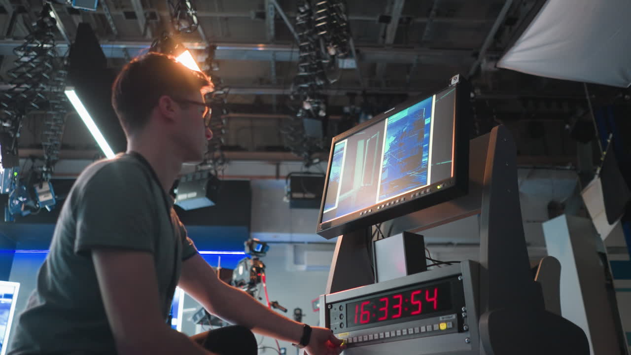 Person adjusting controls on video equipment in TV studio, monitoring multiple screens showing broadcast setup. Timer on control panel displays time, with studio lights and equipment visible