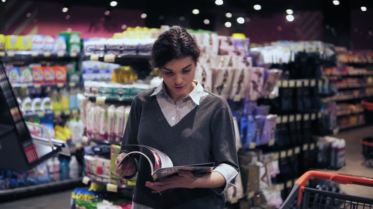 Young beautiful brunette girl at her 20's leafes through magazine and puts it back on shelf trying to decide which one to buy in local supermarket