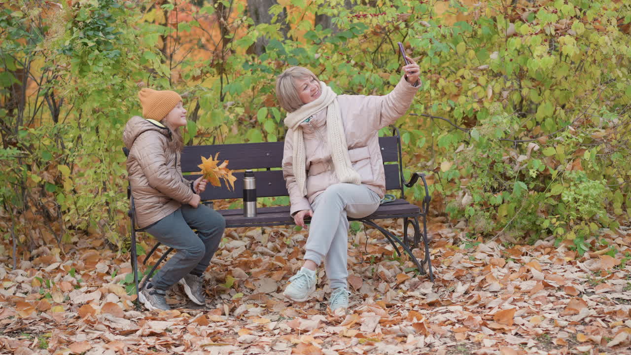 Smiling mother in puffer jacket and scarf sits on park bench taking selfie with smartphone, while daughter in beanie and coat holds yellow leaves, both surrounded by autumn foliage and dry leaves