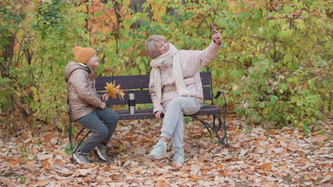 smiling mother in puffer jacket and scarf sits on park bench taking selfie with smartphone, while daughter in beanie and coat holds yellow leaves, both surrounded by autumn foliage and dry leaves