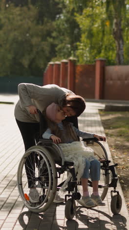 redhead woman leans to kiss cheek of daughter in wheelchair on stone road. little girl with disability enjoys walking in city park with caring mother