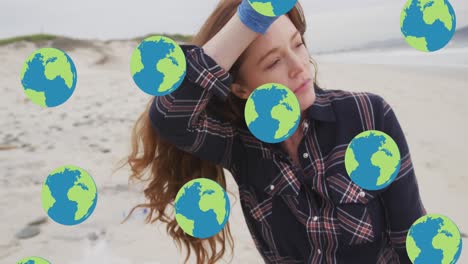 Animation-of-falling-globes-over-caucasian-woman-picking-up-rubbish-from-beach