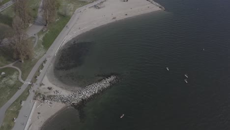 Aerial-fly-by-over-English-Bay-Sunset-Beach-birds-eye-view-of-paddle-board-surfers-enjoying-an-entertaining-summer-experience-of-physical-fitness-while-people-excercise-on-the-seawall