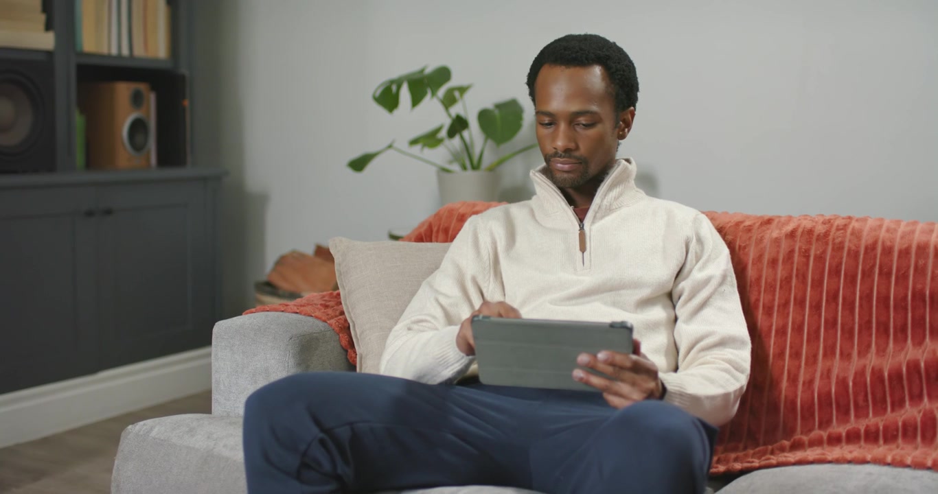 African American man placing tablet on lap, sitting on sofa, tapping, swiping, browsing