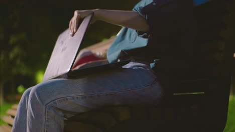 primer plano de una mujer joven sentada al aire libre en un banco por la noche con una bolsa roja cerca, abriendo una computadora portátil, el fondo incluye árboles, un poste de luz y una vegetación exuberante