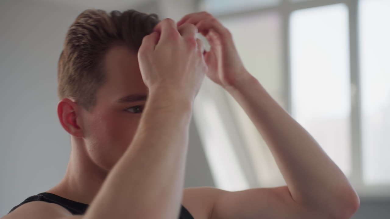 handsome guy in black vest adjusts hair with both hands while studying reflection in large mirror, bright studio space illuminated by daylight through tall window, minimalist interior setting
