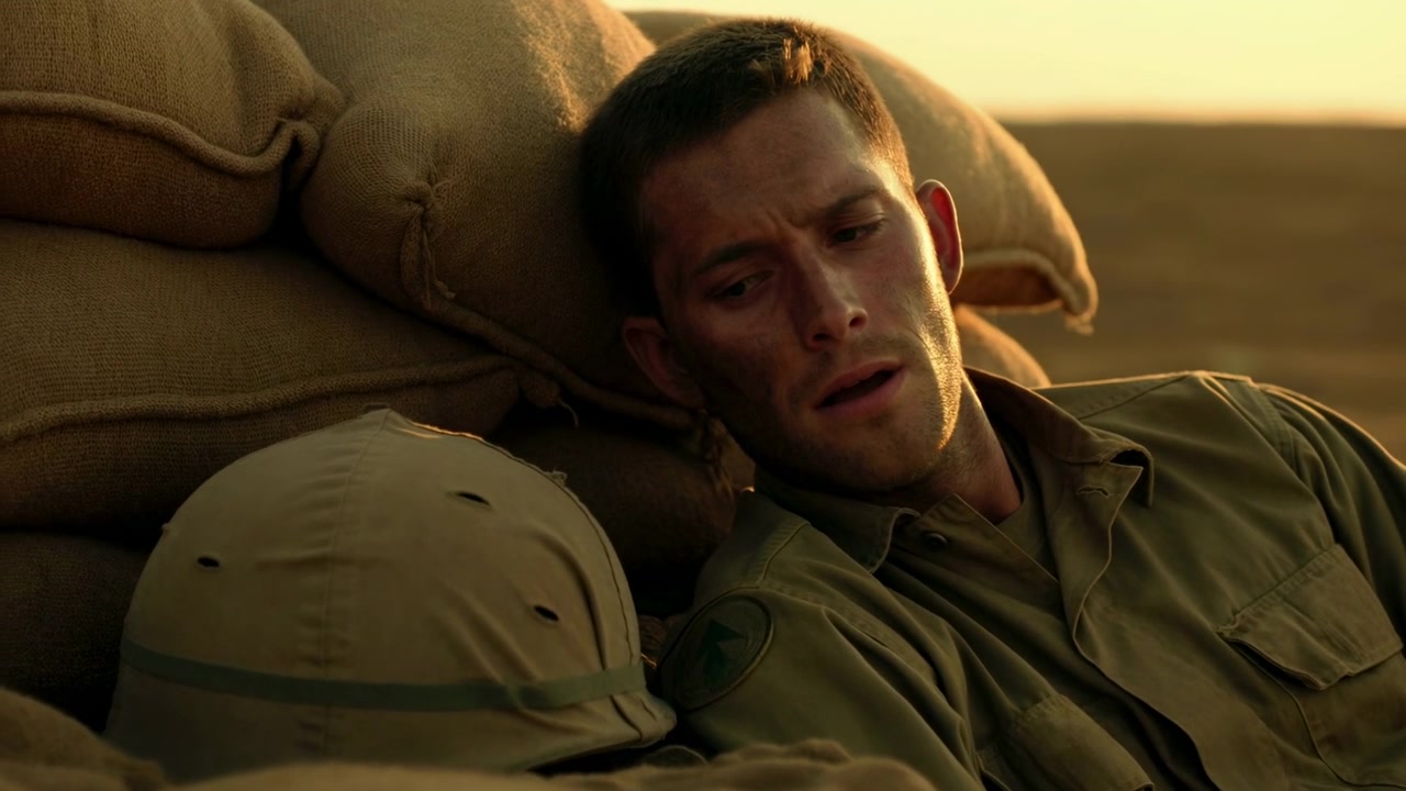 Close cinematic view of male soldier in uniform leaning against burlap sandbags in open desert, turning head toward nearby helmet while resting, with tired expression, dusty textures, warm light