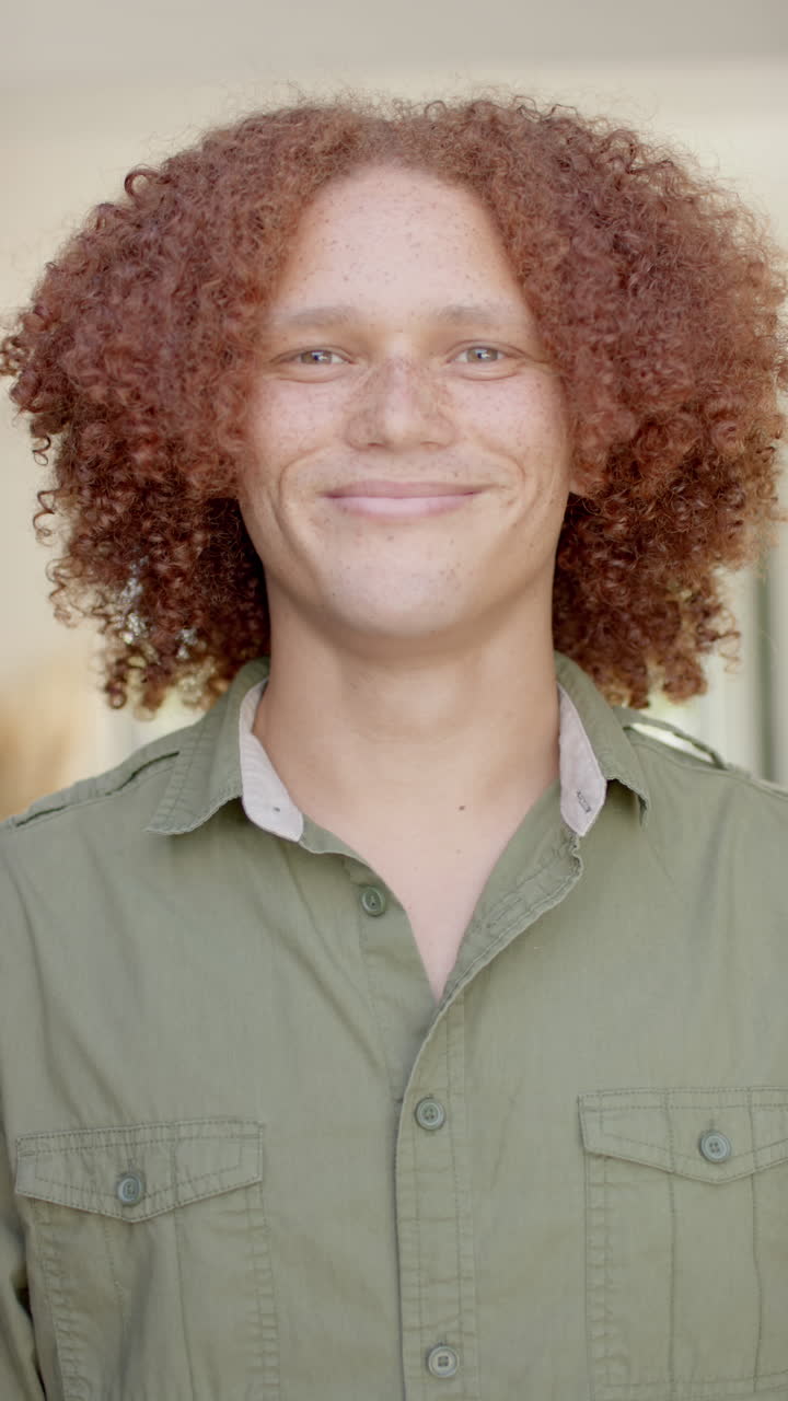 Vertical video of portrait of happy biracial man with curly red hair smiling at home in slow motion