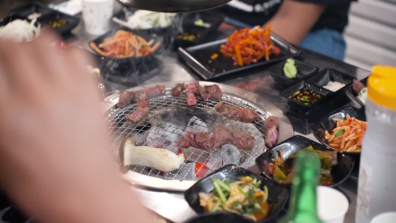 Wide-angle shot of a Korean restaurant tabletop filled with small bowls of colorful side dishes surrounding a central charcoal grill cooking hanu beef and king oyster mushrooms