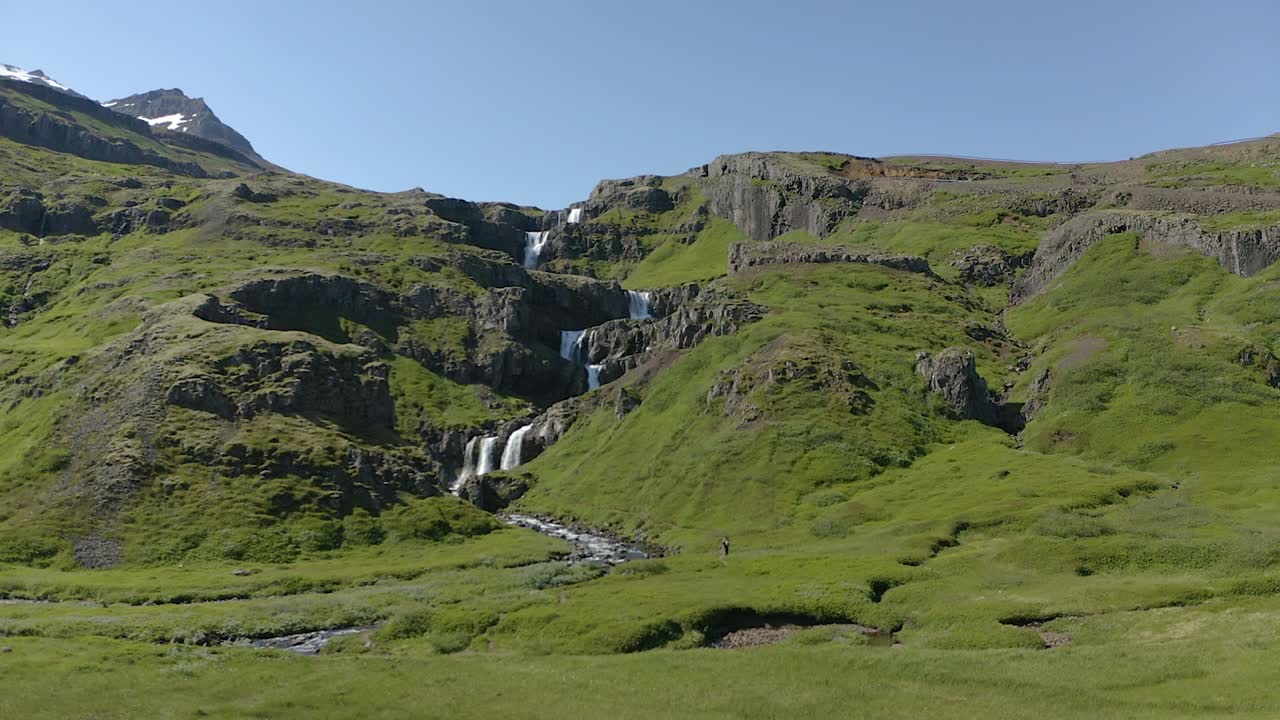Low angle aerial slide up to Klifbrekkufossar waterfall in Iceland running over multiple ledges before arriving on ground level.