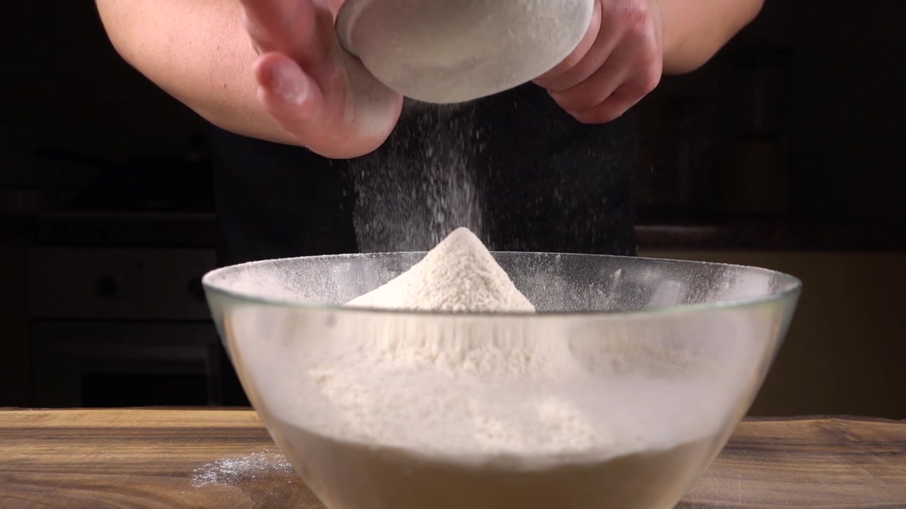 Mengayak tepung menggunakan ayakan ke dalam mangkuk kaca di dapur. Gerakan lambat.