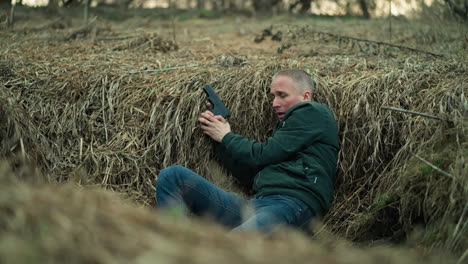 a close view of a man in a green jacket and jeans holding a pistol while lying in a dry, grassy ditch, looking tense scared