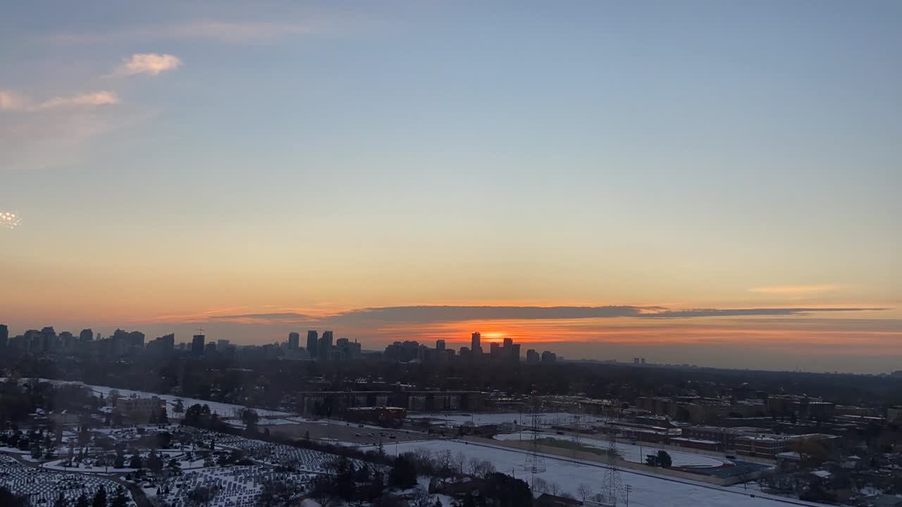 Aerial drone time lapse shows glowing sunset behind urban skyline in Canada with snow-covered ground, soft light, and clear sky capturing peaceful transition from day to evening in winter.
