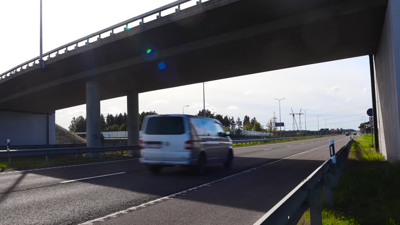 A bridge, overpass or an underpass going over Pärnu highway during summer time while sun is shining and sky is slightly cloudy. Cars and vehicles drive on the asphalt road that has markings on it.