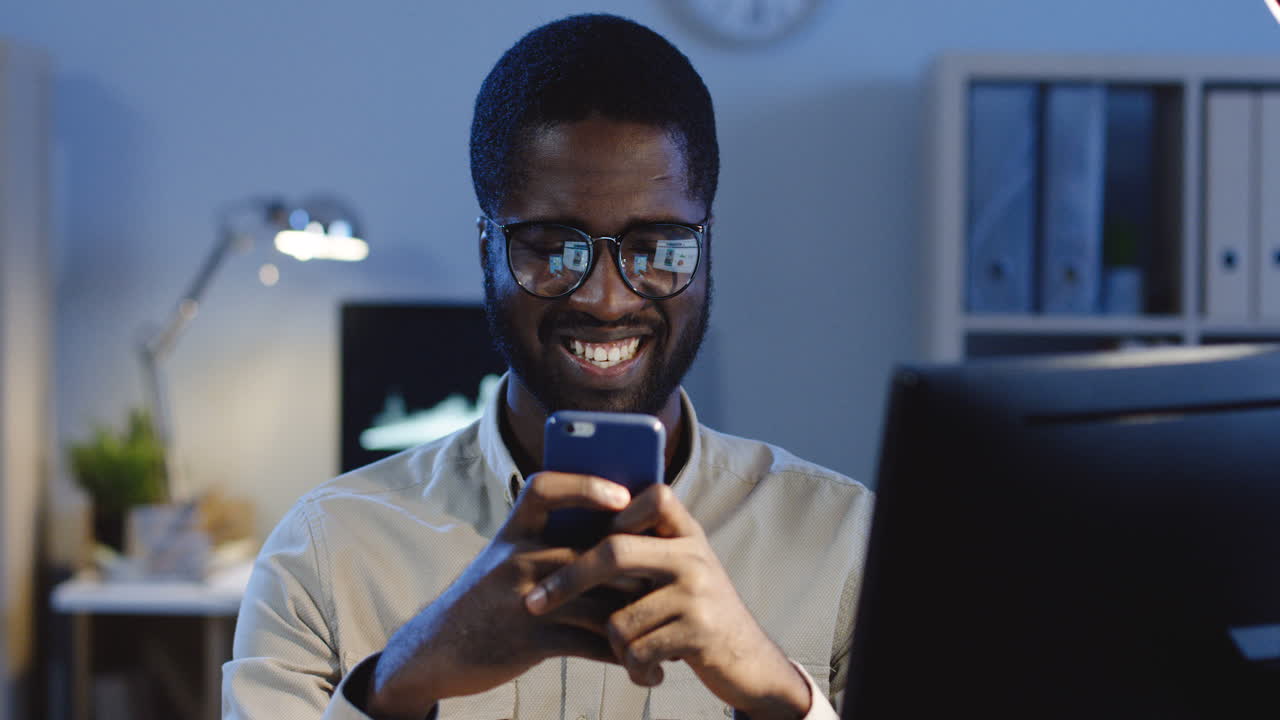 Portrait Of Young Smiled Office Worker Wearing Glasses Typing And Chatting On The Smartphone In The Office At Night