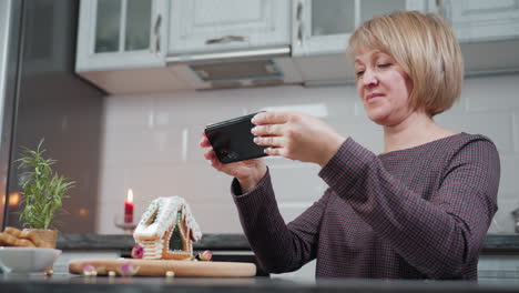 una mujer de mediana edad captura cuidadosamente una foto de su casa de pan de jengibre hecha a mano usando su teléfono inteligente, el fondo presenta una cálida luz de vela, una planta en olla y un ambiente de cocina acogedor