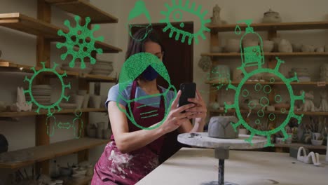 Animation-of-covid-19-cells-and-icons-over-woman-working-in-pottery-studio-wearing-face-mask