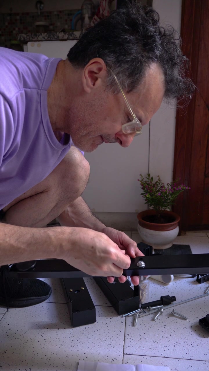 A concentrated middle-aged male holds a paper instruction manual while putting together new furniture on the kitchen floor. Casual lifestyle and home tasks concept.