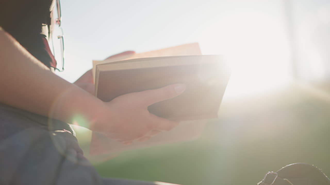 빛은 배경의 흐릿한 초록색에서 빛나고, 밖에서 앉아있는 여성이 들고 있는 책에서 반사되며, 그녀는 검은색 에 안경을 착용하면서 페이지를 부드럽게 뒤집습니다.