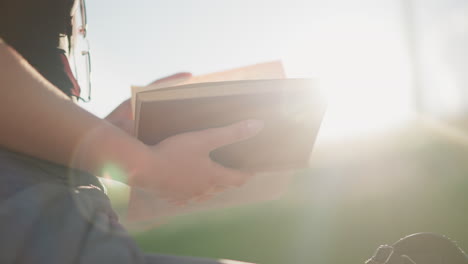 la luz del sol brilla desde la vegetación borrosa en el fondo, reflejándose en el libro sostenido por una mujer sentada al aire libre, ella desliza suavemente las páginas mientras usa gafas en su camiseta negra
