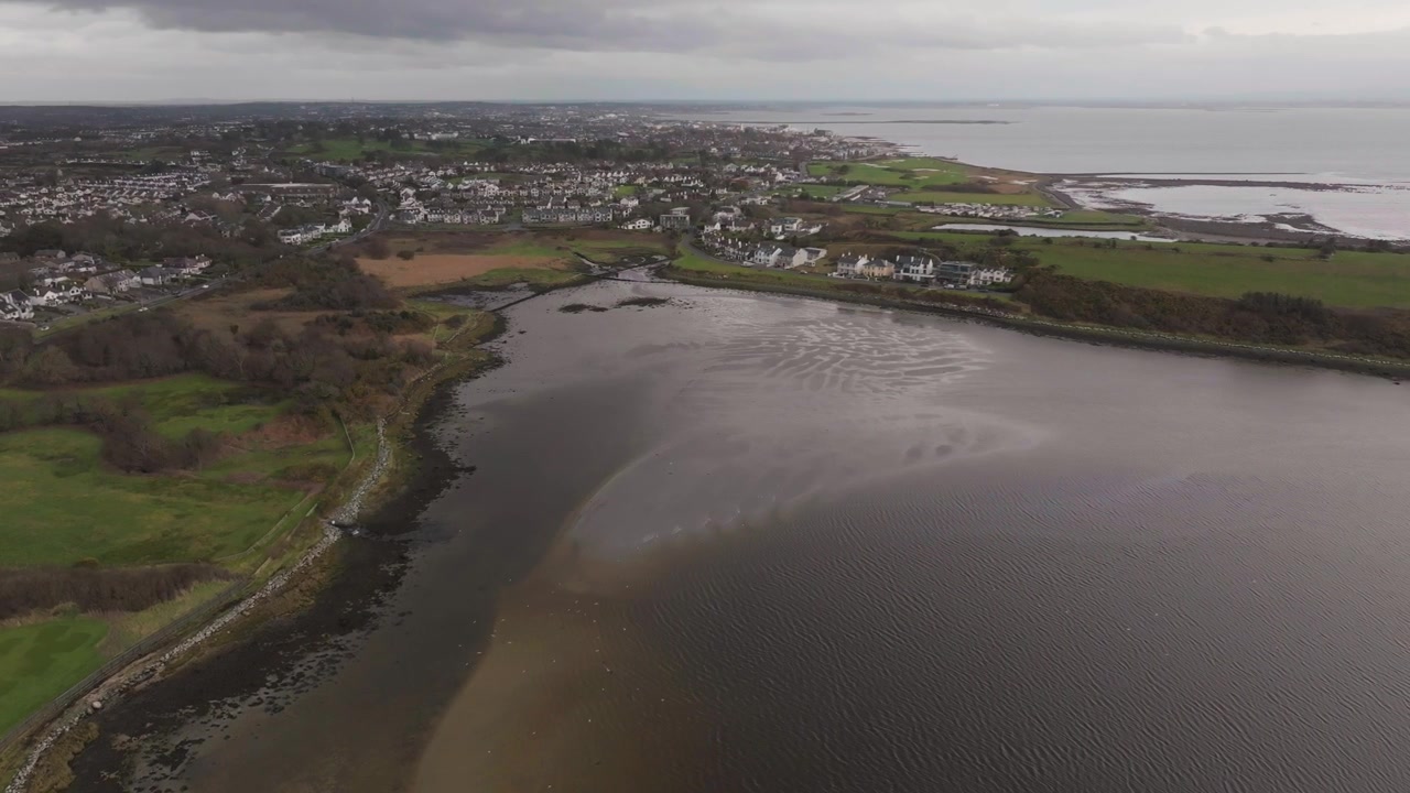 Et filmisk droneoptagelse fra luften, der viser det rolige vand i Rusheen Bay med beboelseslandsbyen Knocknacarra i baggrunden under en overskyet himmel.