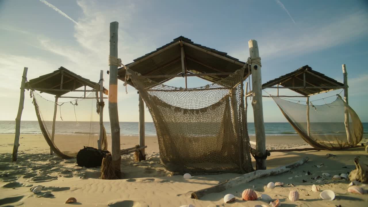 La telecamera arretra per rivelare un baldacchino centrale di paglia e una rete intrecciata che ondeggia su una spiaggia sabbiosa, conchiglie.
