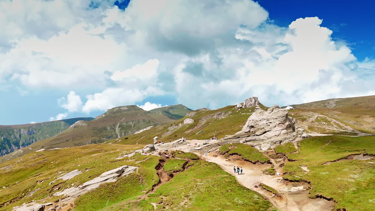 Hikers explore lush green hills at noon. Hikers traverse a winding path through vibrant green hills under a bright blue sky with fluffy clouds during midday