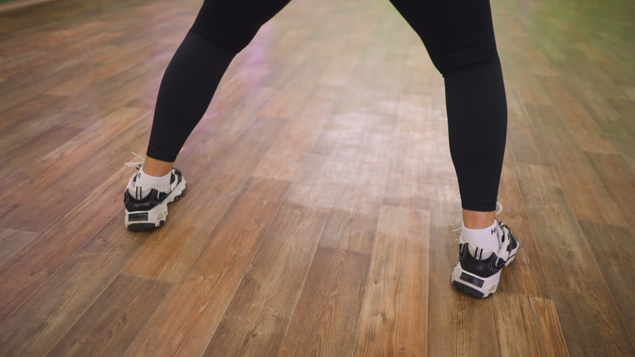 Back view of woman in black leggings and sneakers lifting foot from wooden floor, showing energy and motion during dance workout indoors with strong athletic balance and controlled rhythm