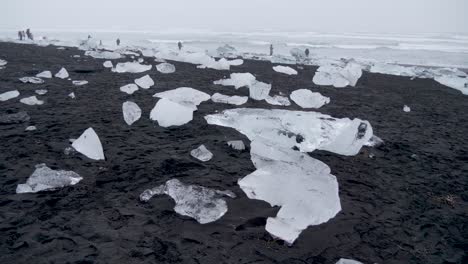 Static,-shot,-of-people-walking-between-ice-blocks-on-a-black-sand-beach-and-the-arctic-sea,-on-a-cloudy-day,-at-Diamond-beach-Islandia,-in-South-coast-of-Iceland