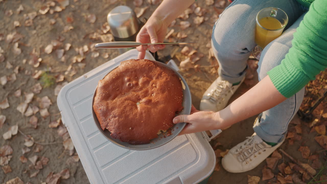 Camping cooler cake held over, camper hands placing golden round cake onto cooler lid, leafy forest floor underfoot, green sweater and jeans visible, glass of orange juice and thermos nearby, warm