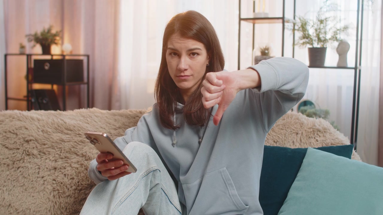Upset woman showing dislike gesture while using smartphone with displeased expression at home