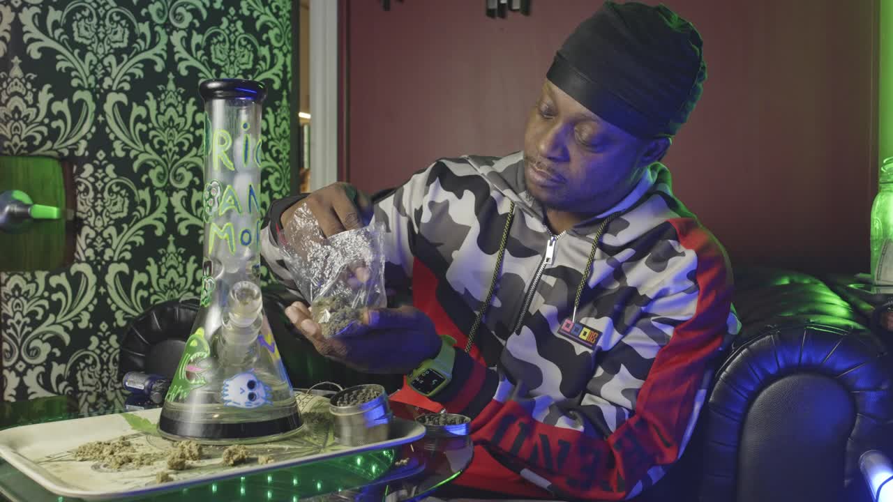 흑인 남자가 마리화나 어리 한 봉투를 들고 새를 맡기 전에 어리를 마리바나 밀러에 어 놓고 테이블 위에 유리 물 을 놓습니다.