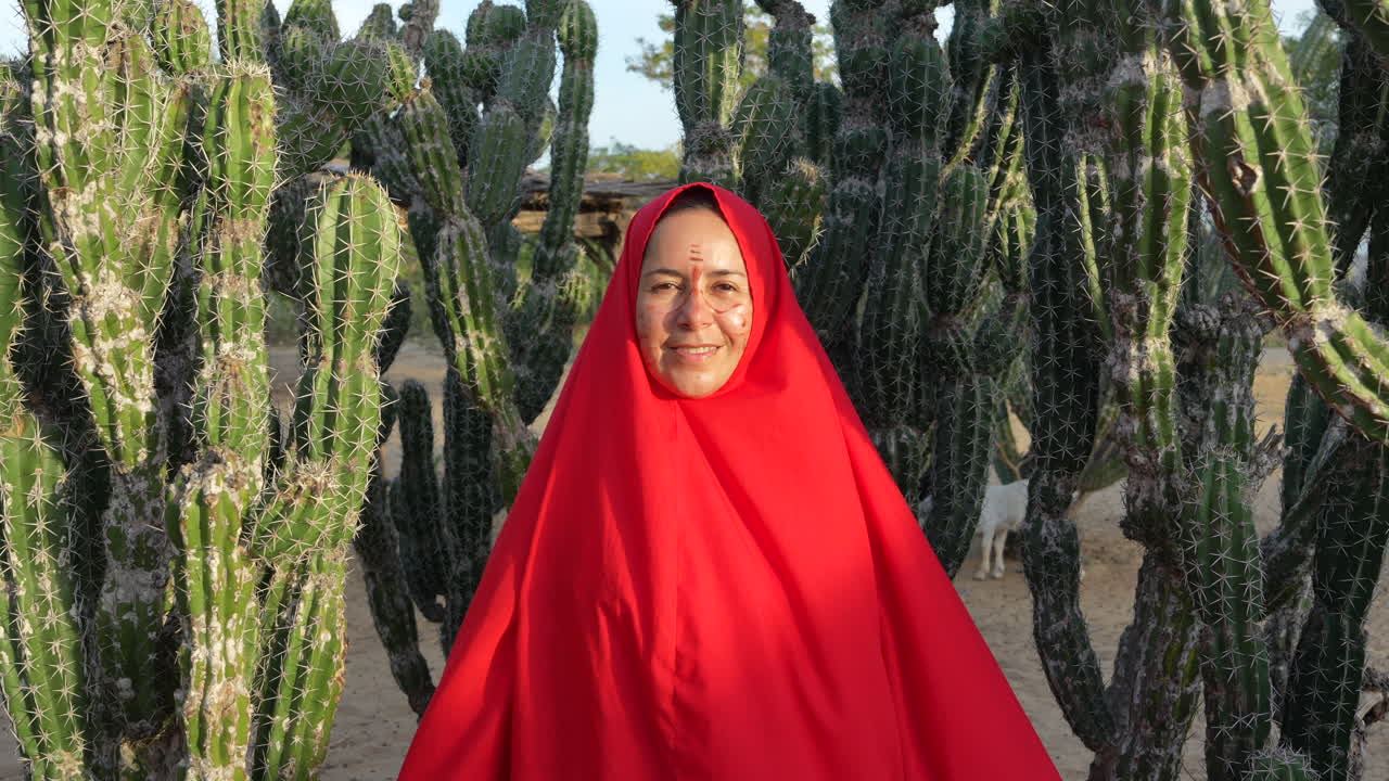 Una mujer Wayuu con un vibrante manto rojo sonríe entre imponentes cactus bajo el cálido sol en La Guajira, Colombia. El paisaje desértico, las texturas nítidas y la iluminación directa crean profundidad cultural