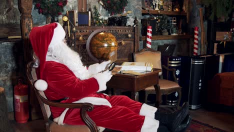 santa is sitting at the table and reading letters. saint nicholas looking at children's letters through a magnifying glass in decorated room at christmas