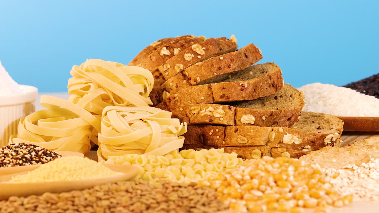 A variety of grains, pasta, and bread arranged against a vibrant blue backdrop. Bright lighting enhances the textures and colors