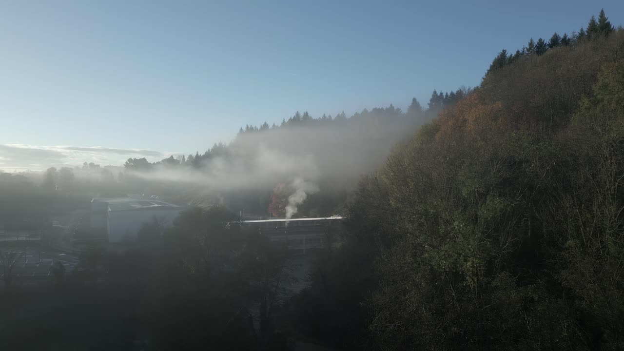 White smoke rises from a factory chimney near a forested hillside, creating a misty layer over sant hilari sacalm in catalonia on a clear morning