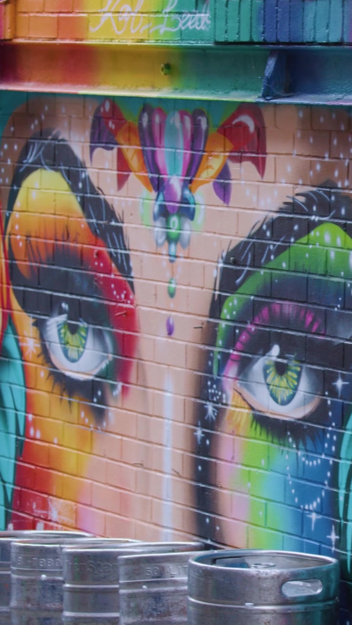 Murale di street art vivido con colori arcobaleno, volti drammatici e barili metallici in un vicolo urbano.